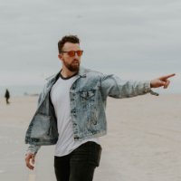 Man in denim jacket and sunglasses pointing on a beach, representing a casual and confident demeanor, aligning with the innovative spirit of the Elite Aeolabs software development team.